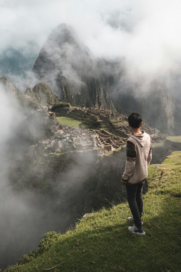 A person standing on a graassy hill, gazing out at the ancient ruins of Machu Picchu, partially obscured by mist and fog. Lima