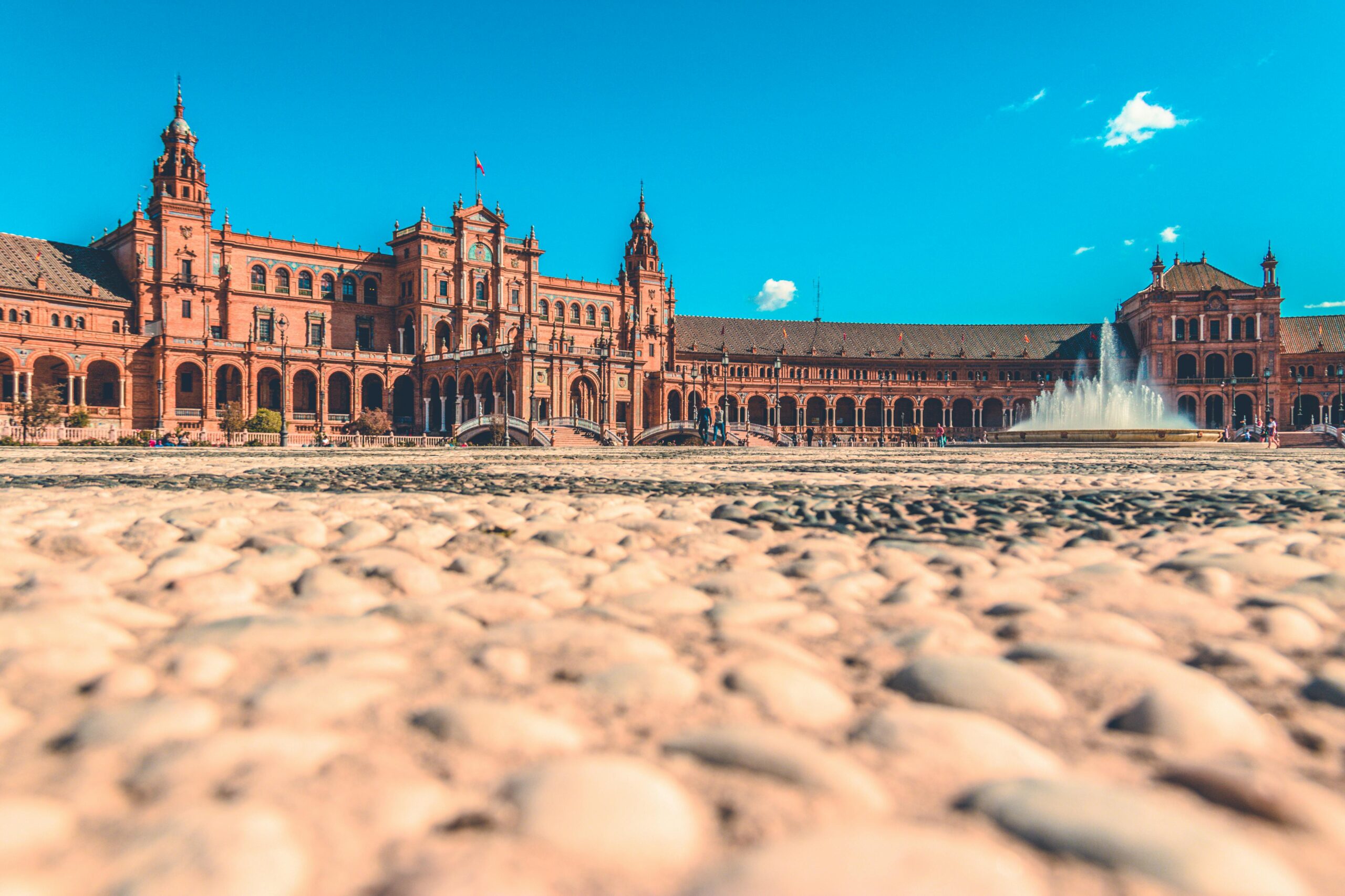 A grand, ornate building with intricate architecture and fountains in the foreground, set against a bright blue sky with clouds, Seville.