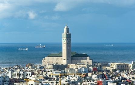 The image shows a large, ornate mosque-like building with a tall minaret tower in the center. The building is surrounded by a dense urban area with other buildings and structures visible in the background. The sky is a bright blue with some clouds. Casablanca