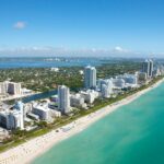 A serene coastal landscape with a rocky shoreline, calm turquoise waters, and a clear blue sky. The scene captures the natural beauty of a quiet beach with scattered rocks and gentle waves. Miami