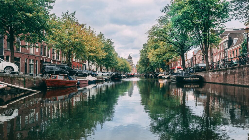 A tranquil canal in Amsterdam, Netherlands, lined with historic brick buildings, boats, and lush trees reflecting in the calm waters.