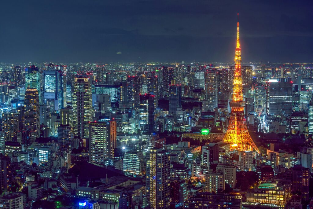 A stunning nighttime cityscape of Tokyo, Japan, featuring the iconic Tokyo Tower illuminated in warm, golden lights against a backdrop of towering skyscrapers and a deep blue sky.