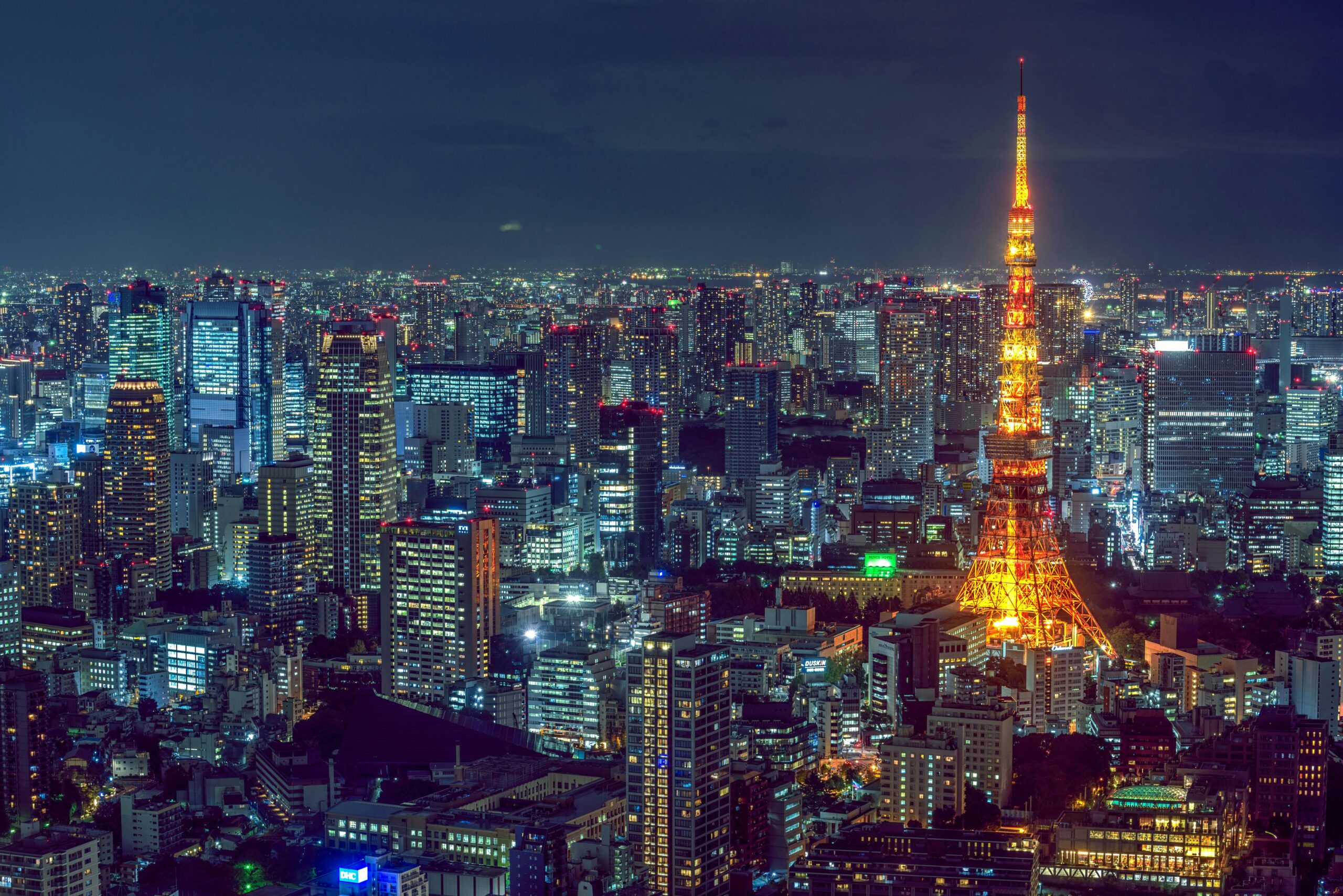 A stunning nighttime cityscape of Tokyo, Japan, featuring the iconic Tokyo Tower illuminated in warm, golden lights against a backdrop of towering skyscrapers and a deep blue sky.